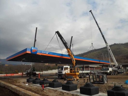 Una gru solleva una grande tettoia blu e arancione per una stazione di servizio in un cantiere edile sotto un cielo nuvoloso.