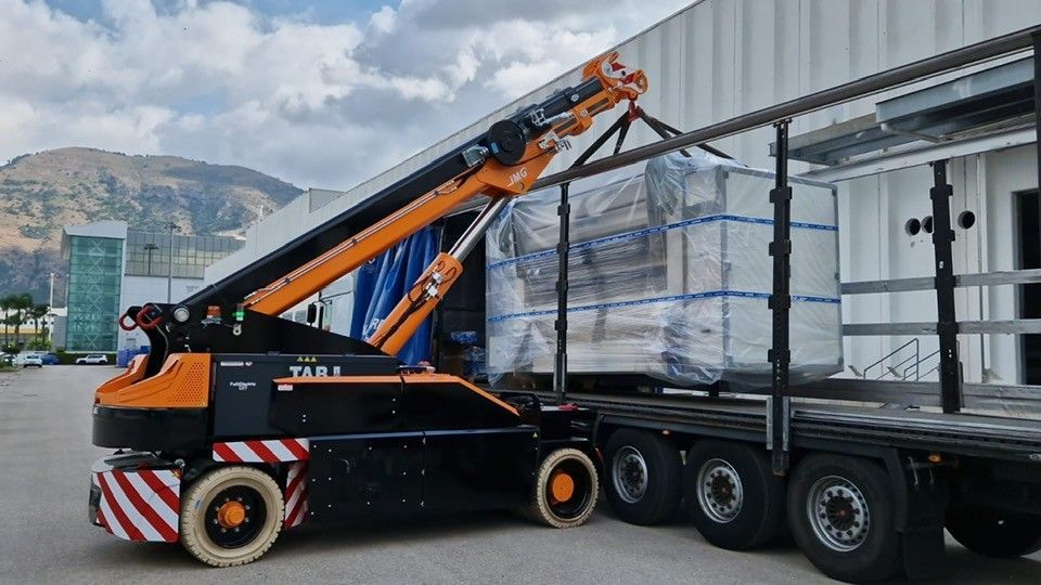 Un carrello elevatore arancione carica pallet imballati su un semirimorchio in un piazzale industriale, con le montagne sullo sfondo.