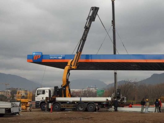 Una gru solleva una lunga trave blu e arancione su un camion in un piazzale industriale.