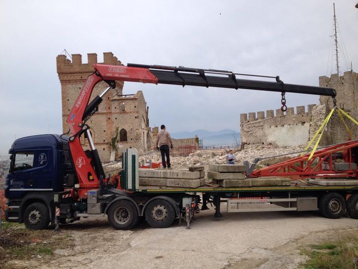 Un camion con gru solleva una capriata rossa accanto a un edificio in pietra in fase di ristrutturazione.
