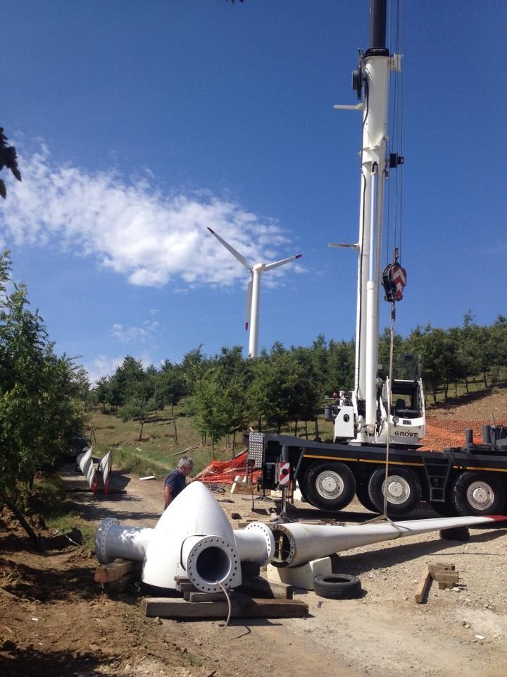 Un camion con gru solleva un componente cilindrico bianco in un cantiere edile sotto un cielo azzurro.
