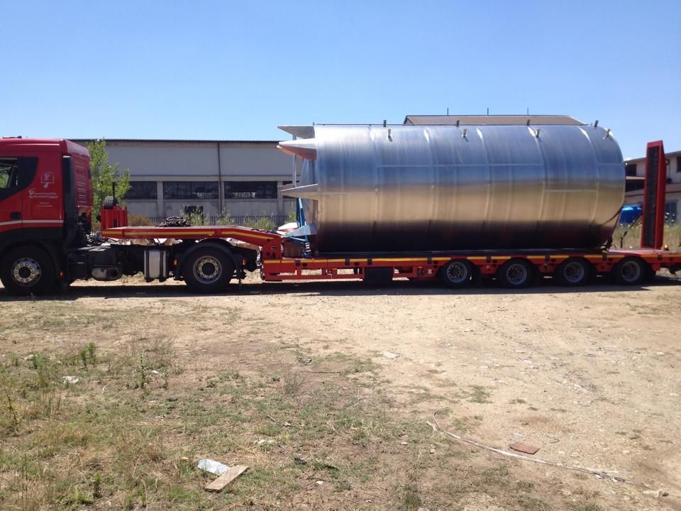 Un camion rosso trasporta un grande serbatoio cilindrico argentato su un rimorchio a pianale all'aperto.