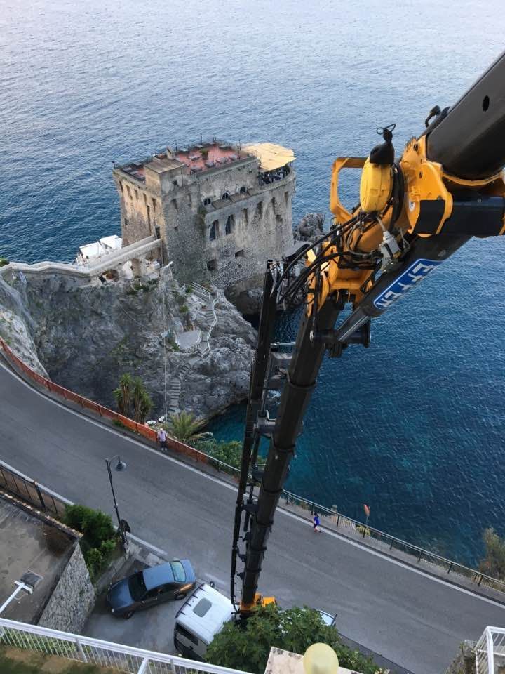 Una gru da costruzione ispeziona una torre sul lungomare, su una scogliera rocciosa sopra una strada e l'acqua blu.