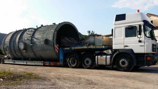 Un camion bianco che trasporta un enorme serbatoio cilindrico di metallo su un rimorchio a pianale all'aperto.