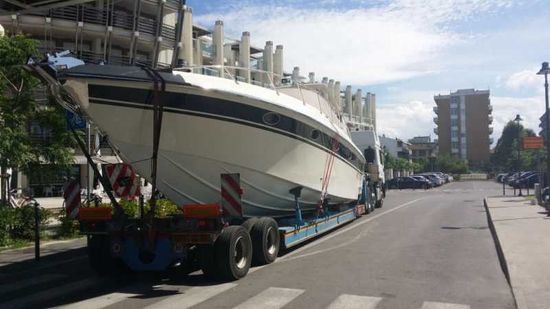 Una grande imbarcazione bianca su un rimorchio viene trainata lungo una strada cittadina vicino al lungomare.