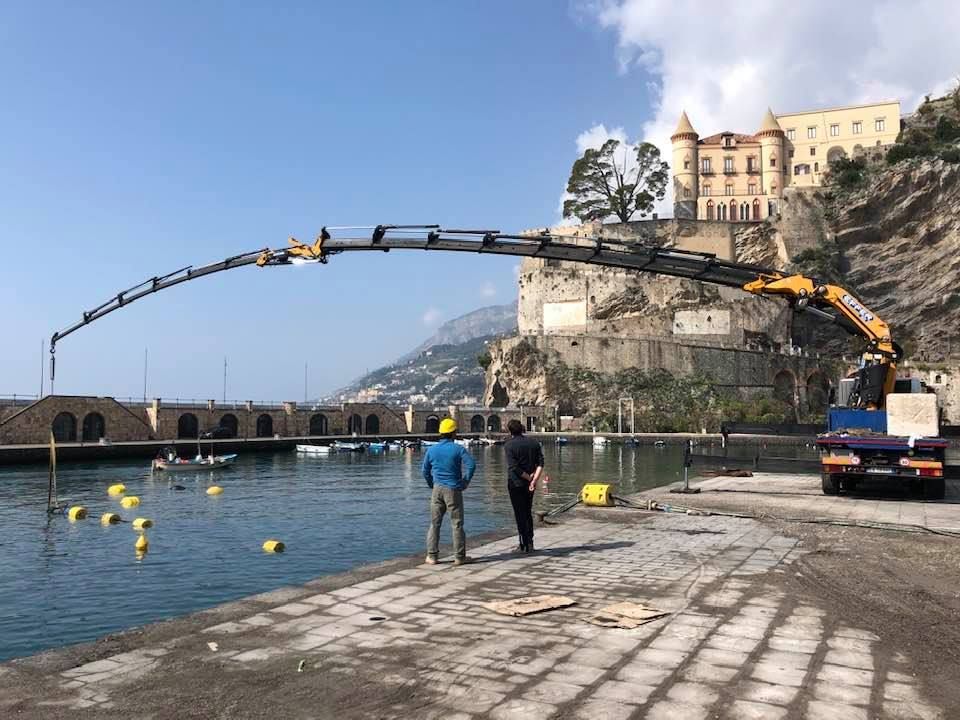 Una gru galleggiante solleva una lunga struttura ad arco sopra un porto vicino a un castello su una scogliera.