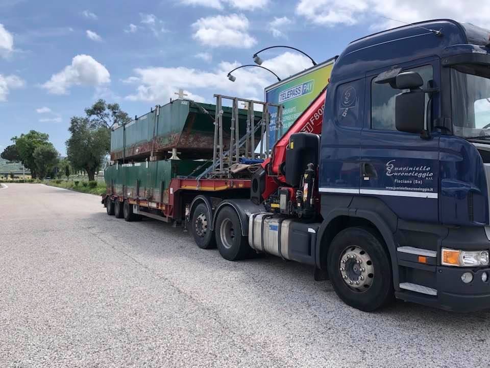 Un camion blu che trasporta rimorchi verdi impilati su una strada sotto un cielo nuvoloso.