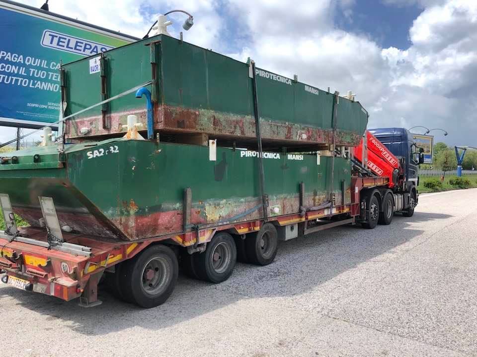 Un grande cassonetto industriale verde su un camion a pianale rosso parcheggiato su una strada sotto un cielo nuvoloso.