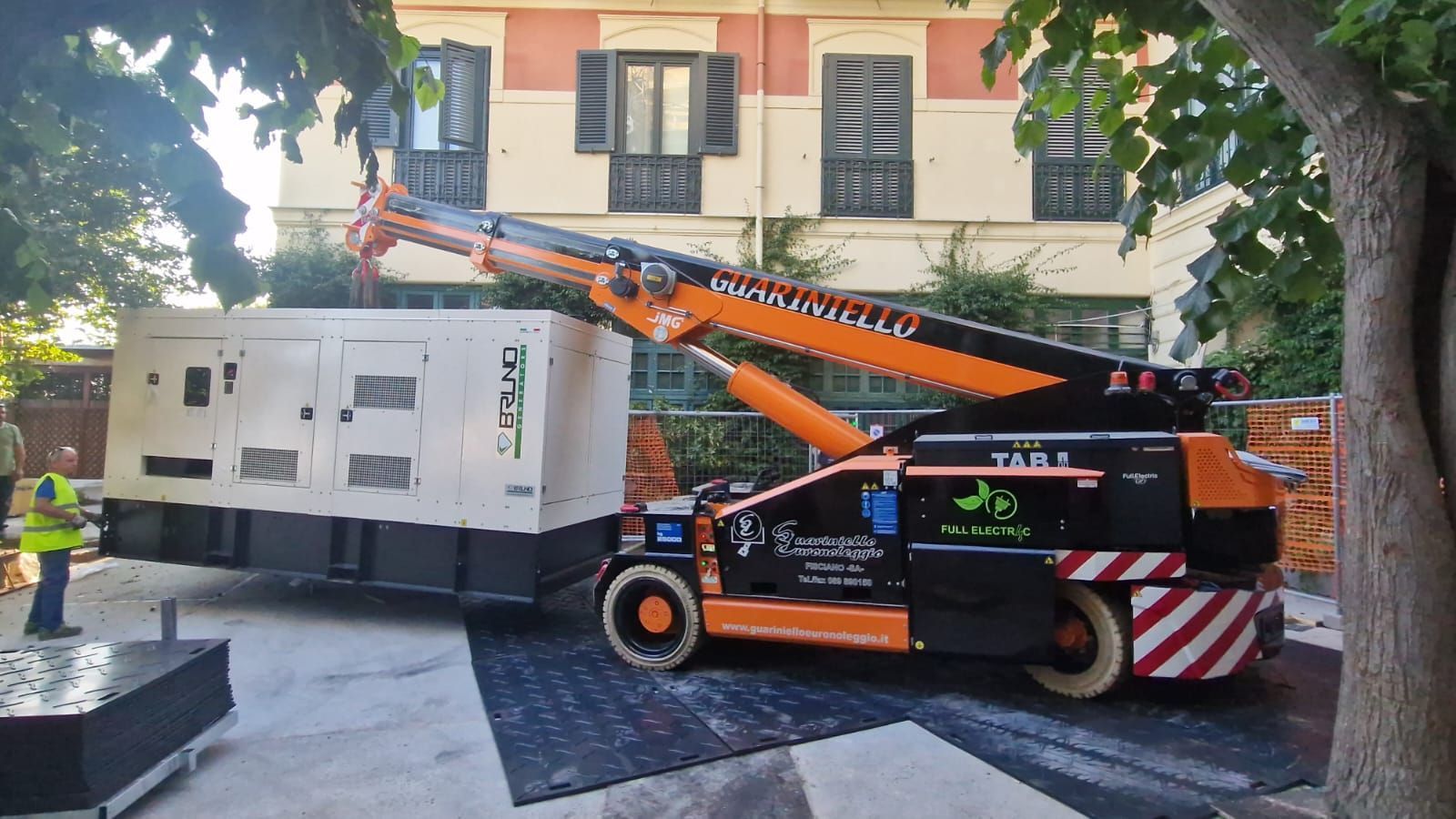 Un carro attrezzi solleva un furgone bianco su una strada cittadina, accanto a un edificio e ad alcuni alberi.