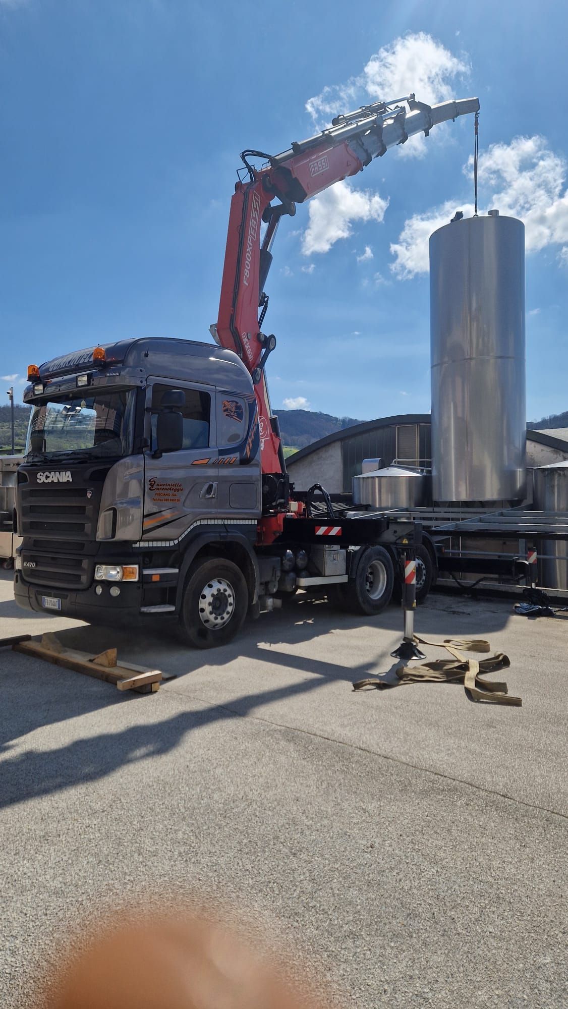 Un camion nero con una gru rossa solleva un grande cilindro metallico all'aperto, sotto un cielo azzurro.