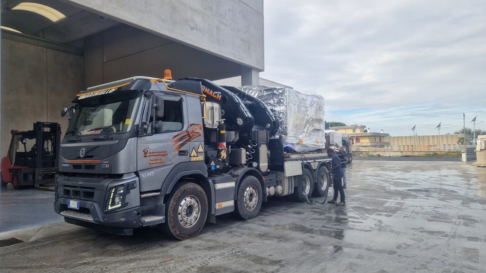 Un camion grigio per carichi pesanti con gru è parcheggiato sotto una struttura in cemento accanto a un carico imballato in un piazzale bagnato.