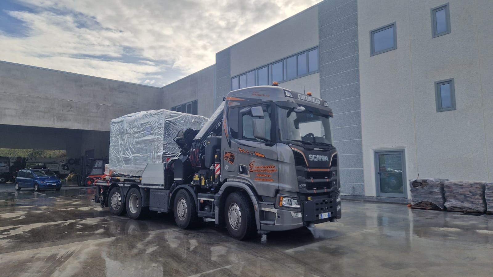 Un camion a pianale scarica un pallet imballato all'esterno di un magazzino in un'area industriale umida.