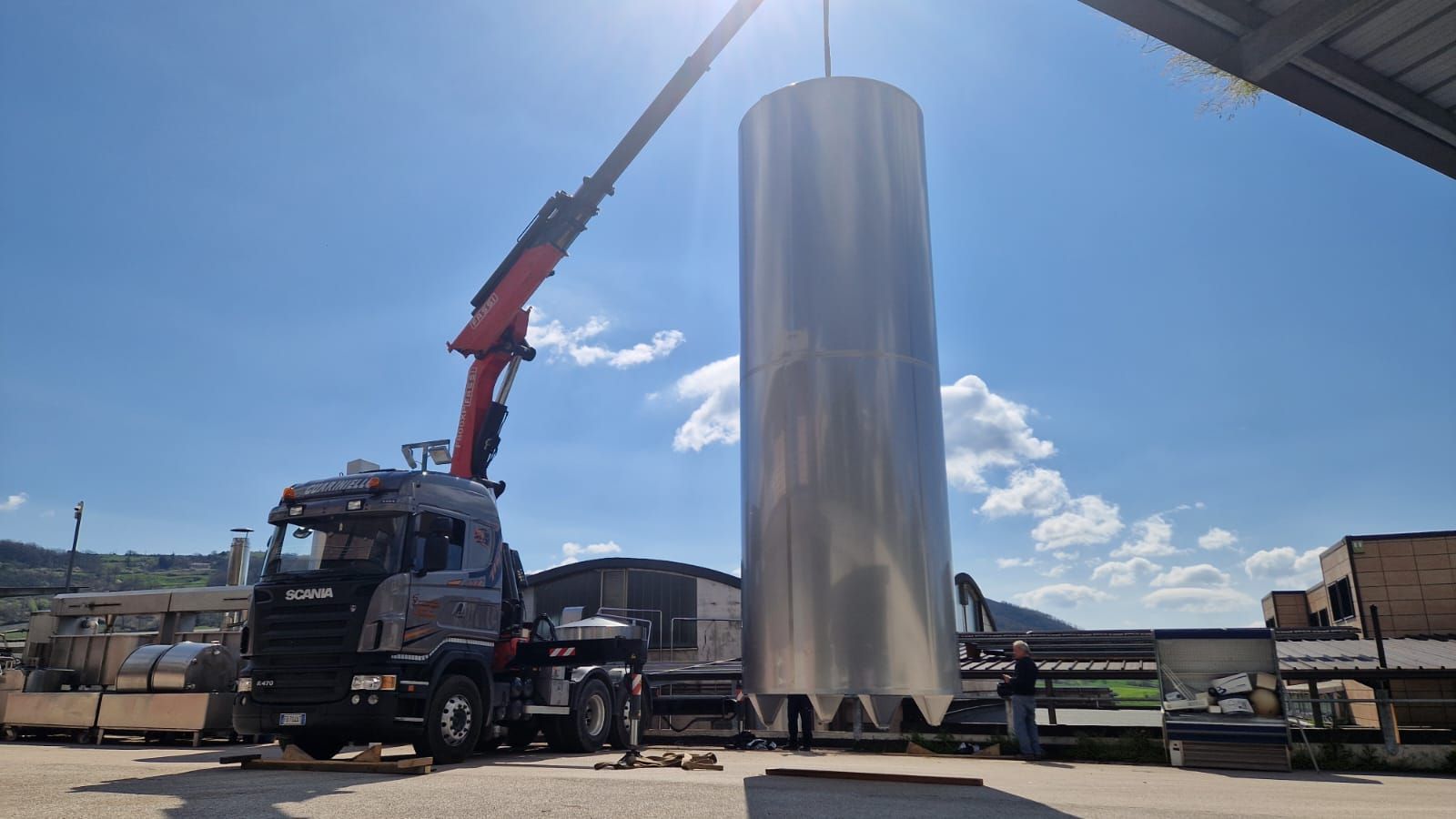 Un camion con gru rosso solleva un alto serbatoio cilindrico argentato in un piazzale industriale sotto un cielo azzurro.