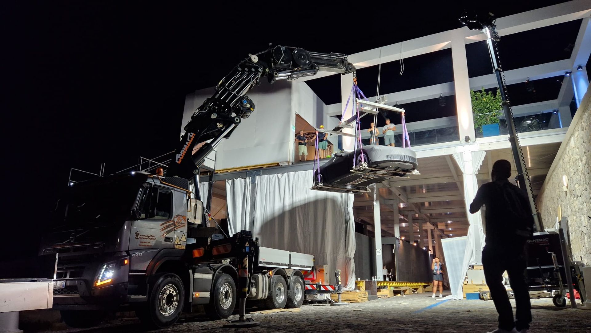 Di notte, una gru solleva una struttura modulare bianca, con operai e un camion sul posto.