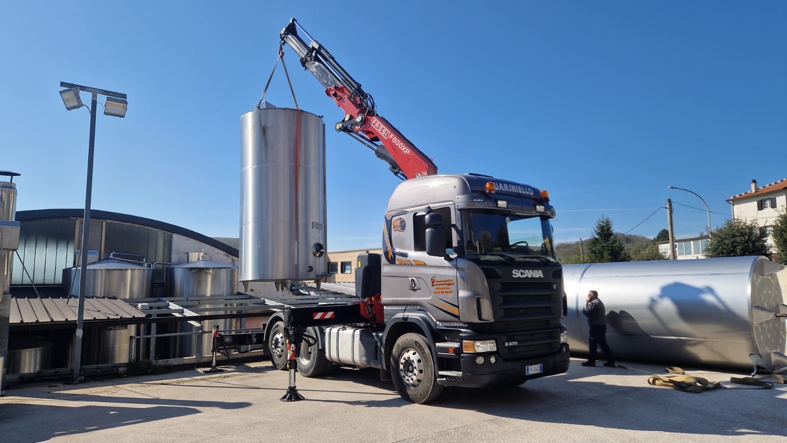 Un'autocisterna argentata con gru rossa carica un grande serbatoio metallico verticale in un sito industriale all'aperto.