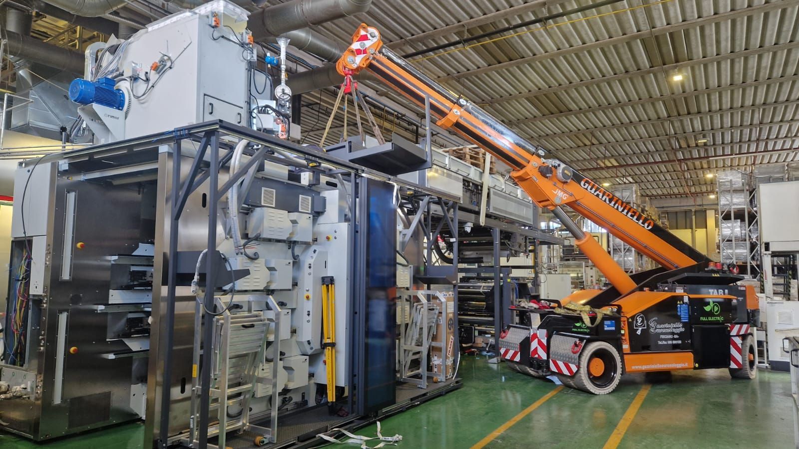 Gru arancione posizionata accanto a un grande macchinario industriale all'interno di un magazzino di una fabbrica.
