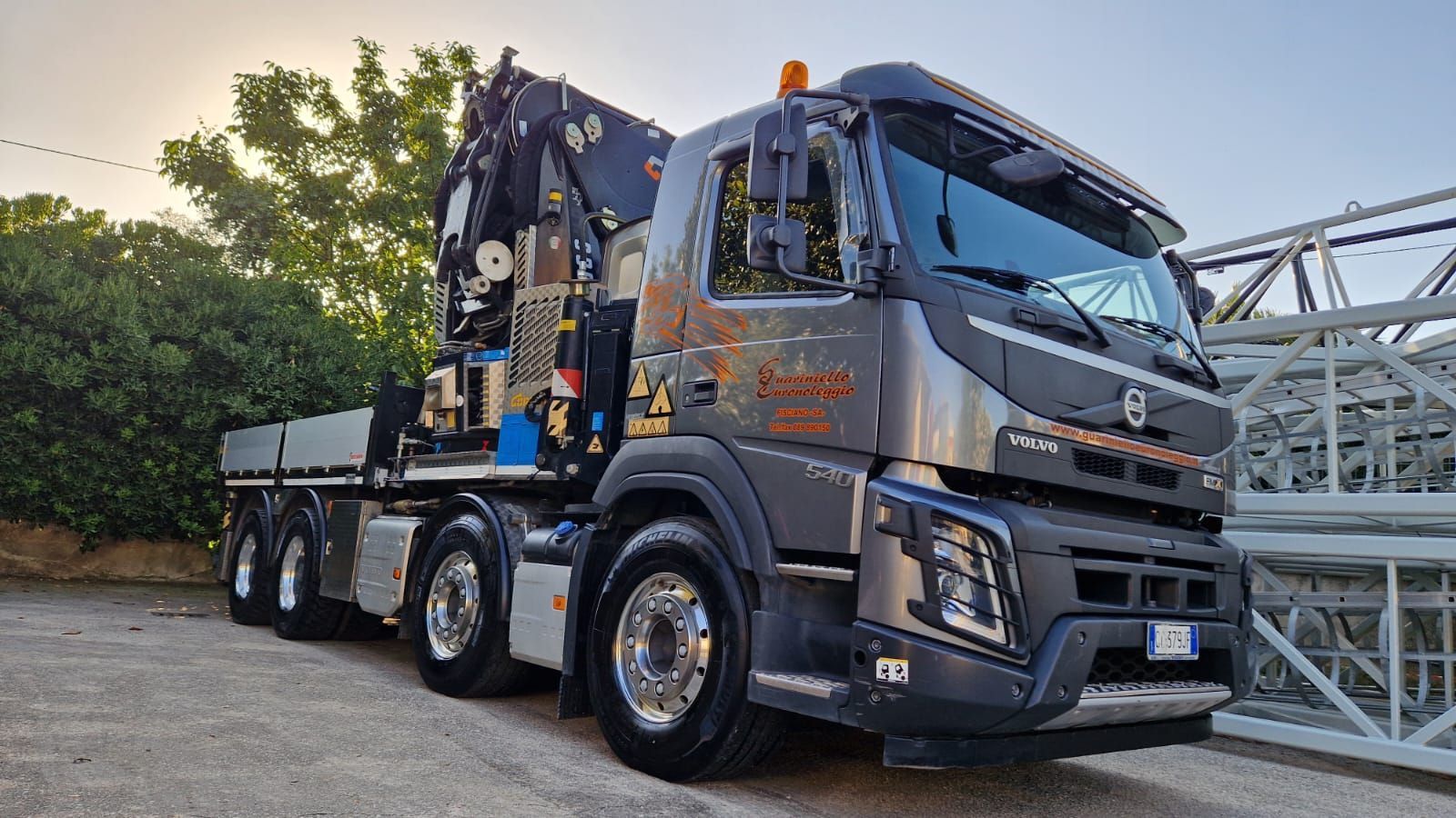 Un camion gru grigio scuro parcheggiato all'aperto accanto a una struttura reticolare metallica al tramonto.