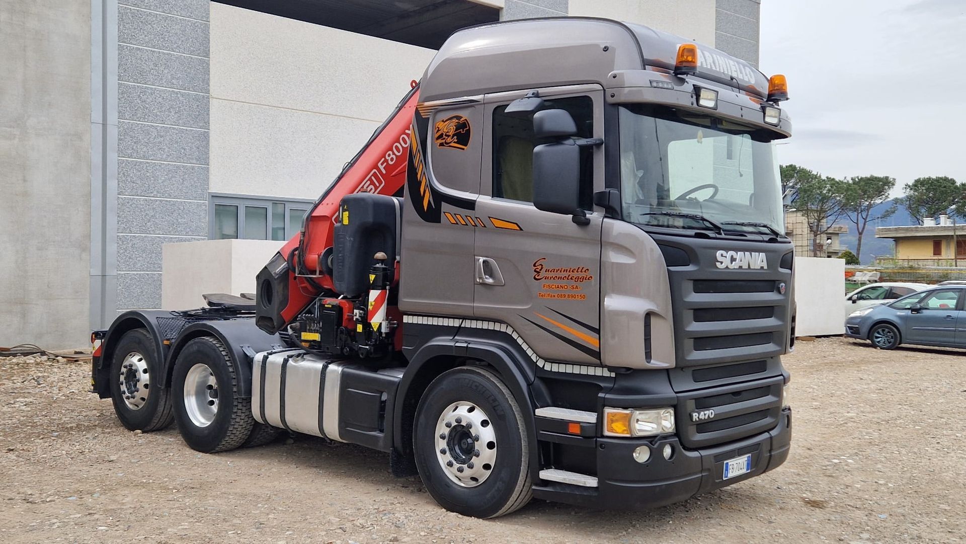 Un semirimorchio Scania grigio con gru rossa è parcheggiato accanto a un edificio su un piazzale ghiaioso.