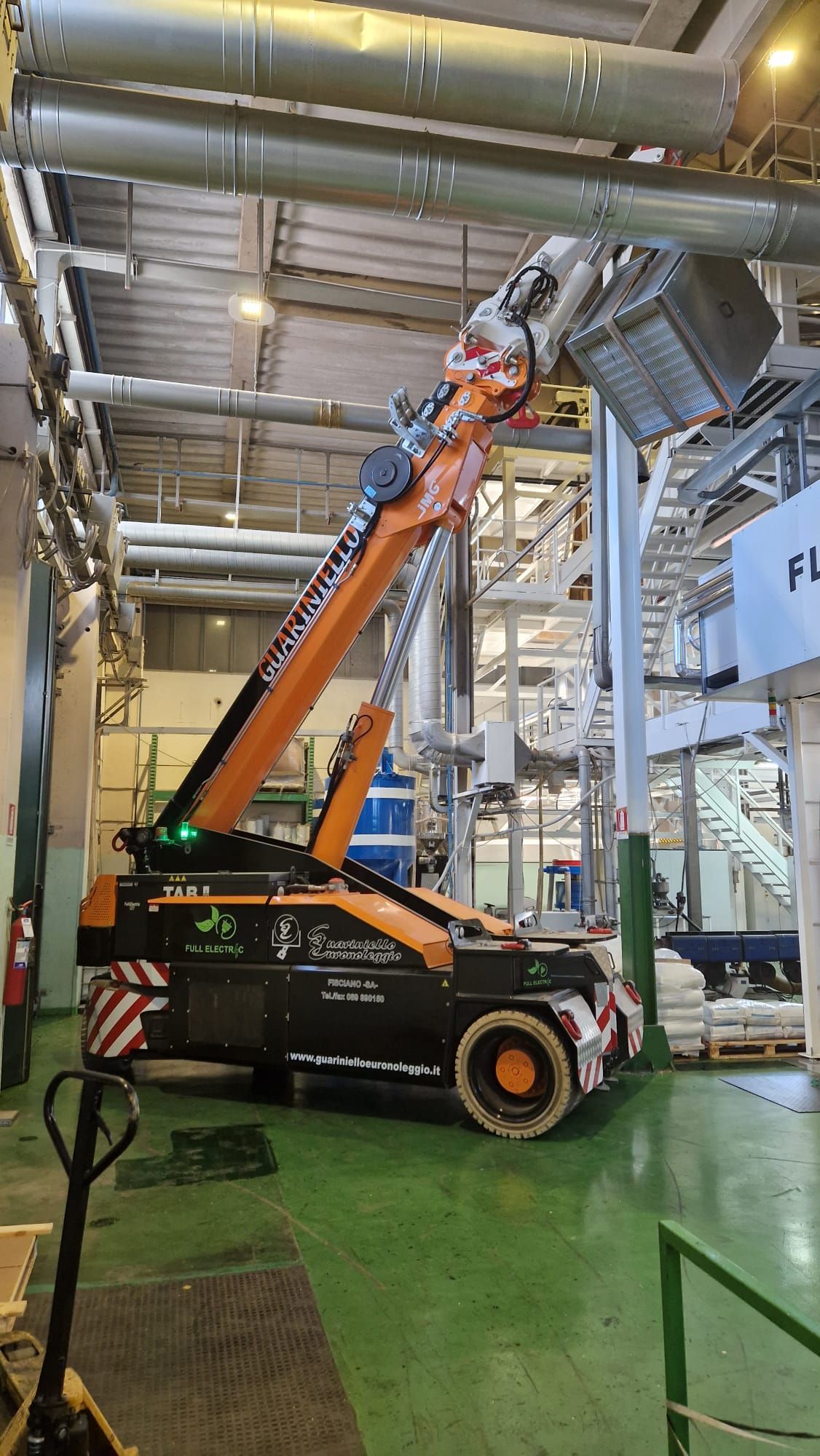Un carrello elevatore arancione solleva un cestello per lavoratori in un magazzino industriale sotto grandi tubature aeree.