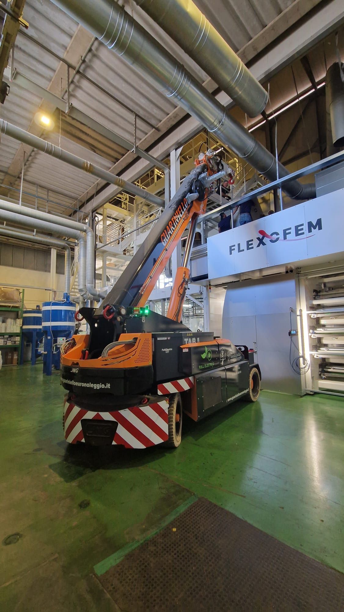 Una piattaforma aerea arancione in un magazzino industriale, che si protende verso le tubature aeree vicino a un'insegna FlexForm.