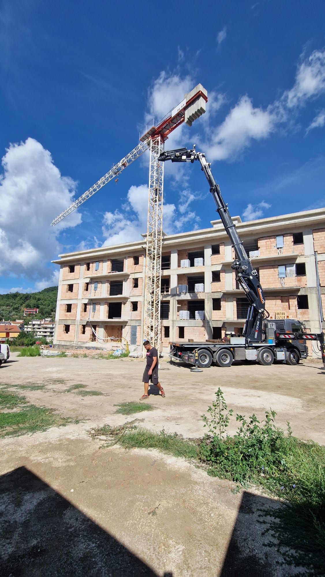 Una gru da cantiere solleva materiali accanto a un condominio beige sotto un cielo azzurro.