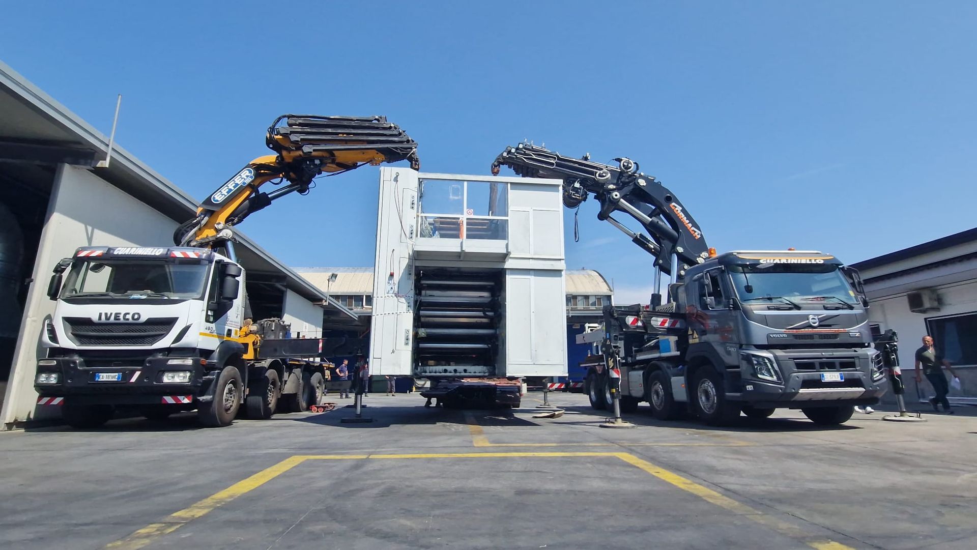 Due camion con gru estensibili sollevano una grande struttura bianca in un'area industriale.