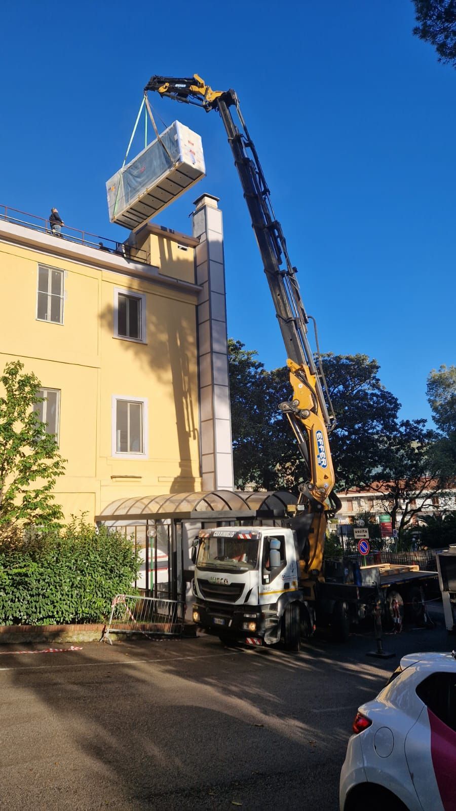 Una gru solleva un container sul tetto di un condominio giallo in una giornata di sole.