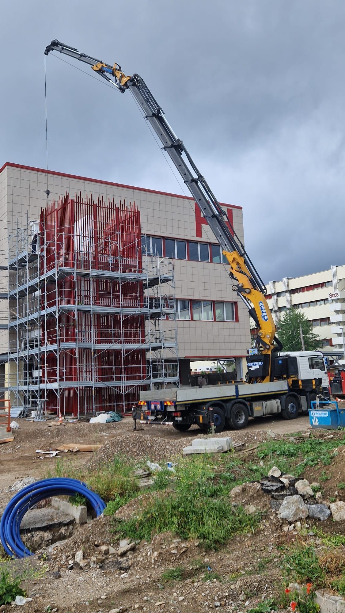 Camion da cantiere con lungo braccio della gru che solleva materiali accanto a un cantiere parzialmente coperto sotto un cielo grigio