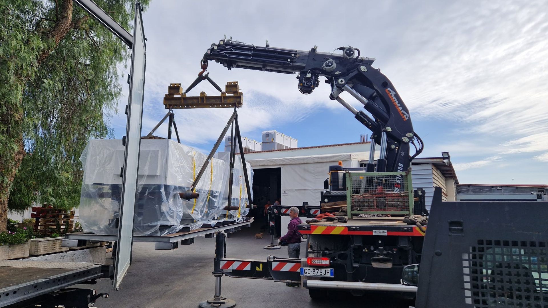 Un camion con gru solleva un oggetto avvolto con cinghie su una strada accanto a una casa.