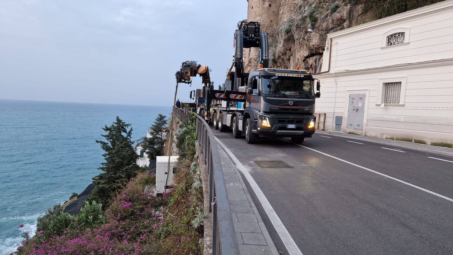 Un camion con gru su una strada costiera a picco sul mare e sulle scogliere, con un altro camion davanti.