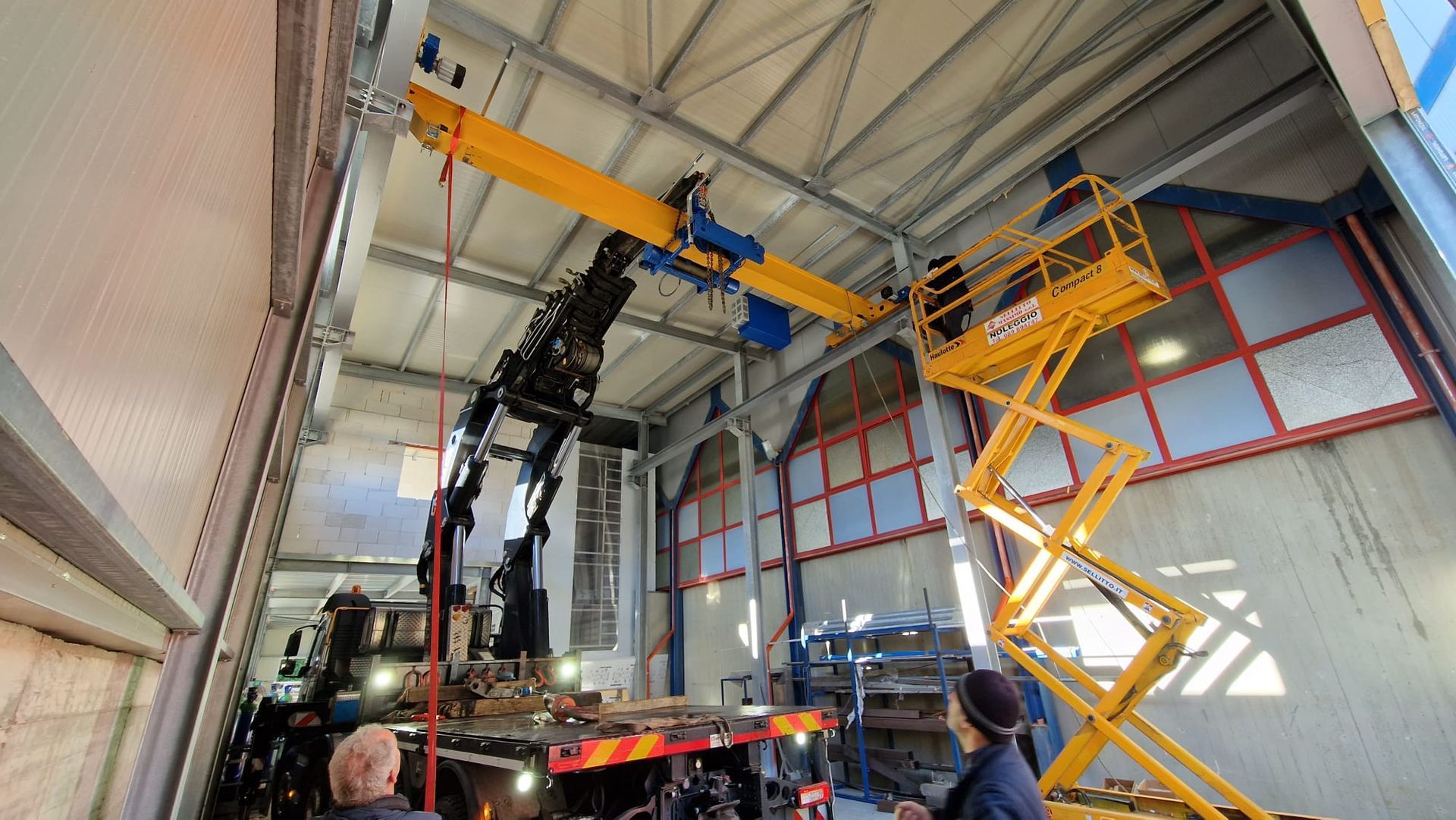 Piattaforma elevatrice industriale al servizio di un magazzino con soffitto alto, composta da una piattaforma a forbice gialla e un braccio di gru nero posizionato vicino al tetto.