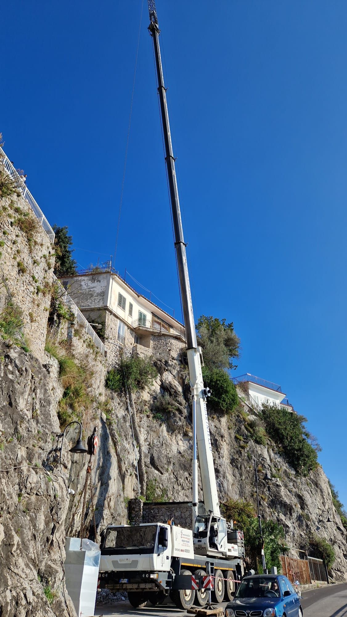 Una gru solleva un carico accanto a una ripida scogliera sul mare, con edifici sovrastanti, sotto un cielo azzurro e limpido.