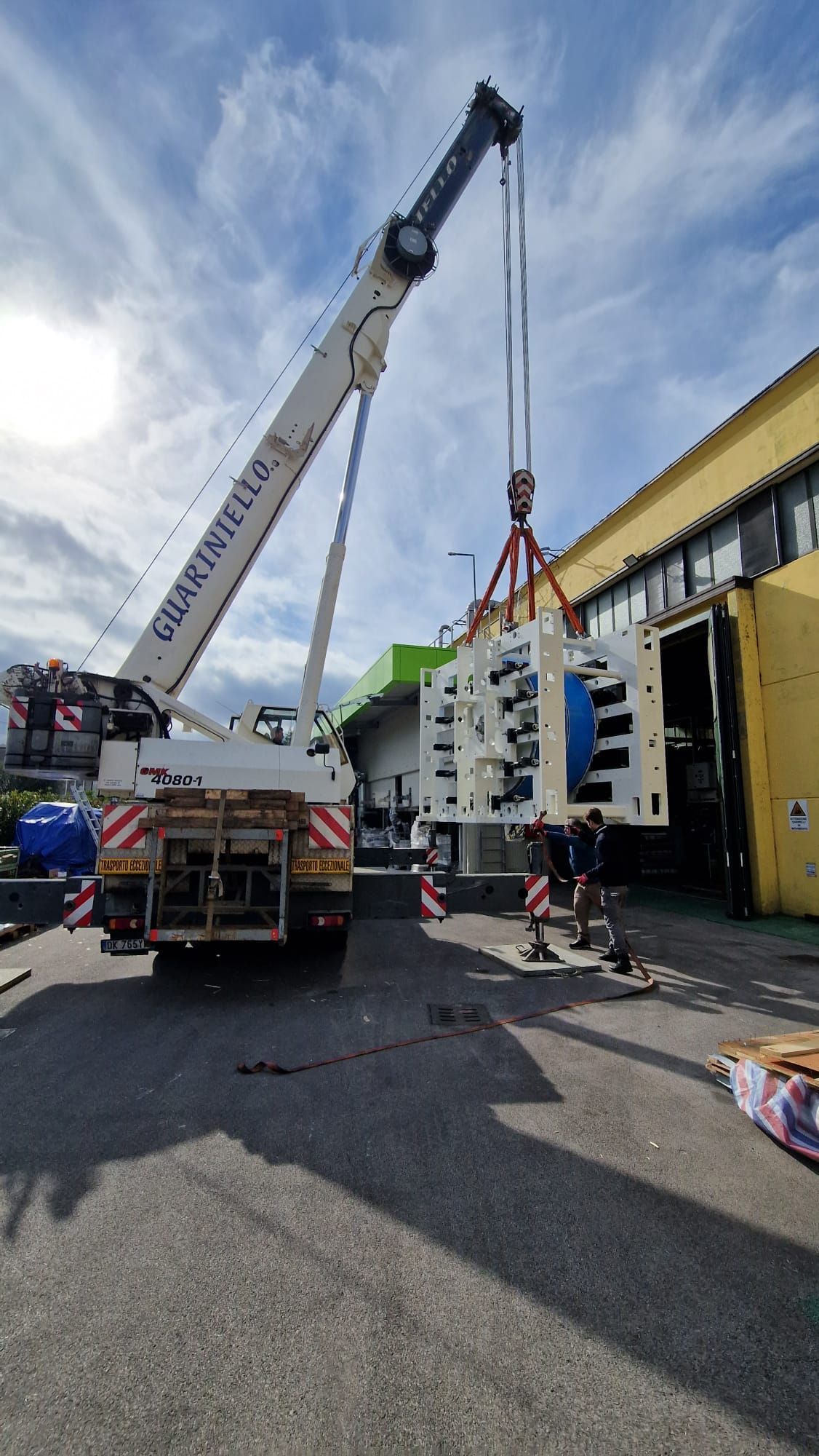 Una gru solleva un modulo prefabbricato bianco accanto a un edificio giallo su una strada.