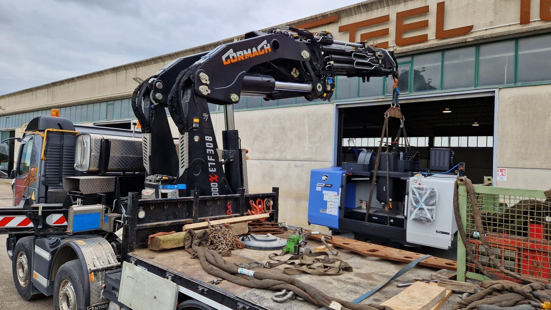 Un escavatore nero carica detriti su un camion accanto a un cantiere per la costruzione di una struttura in acciaio.