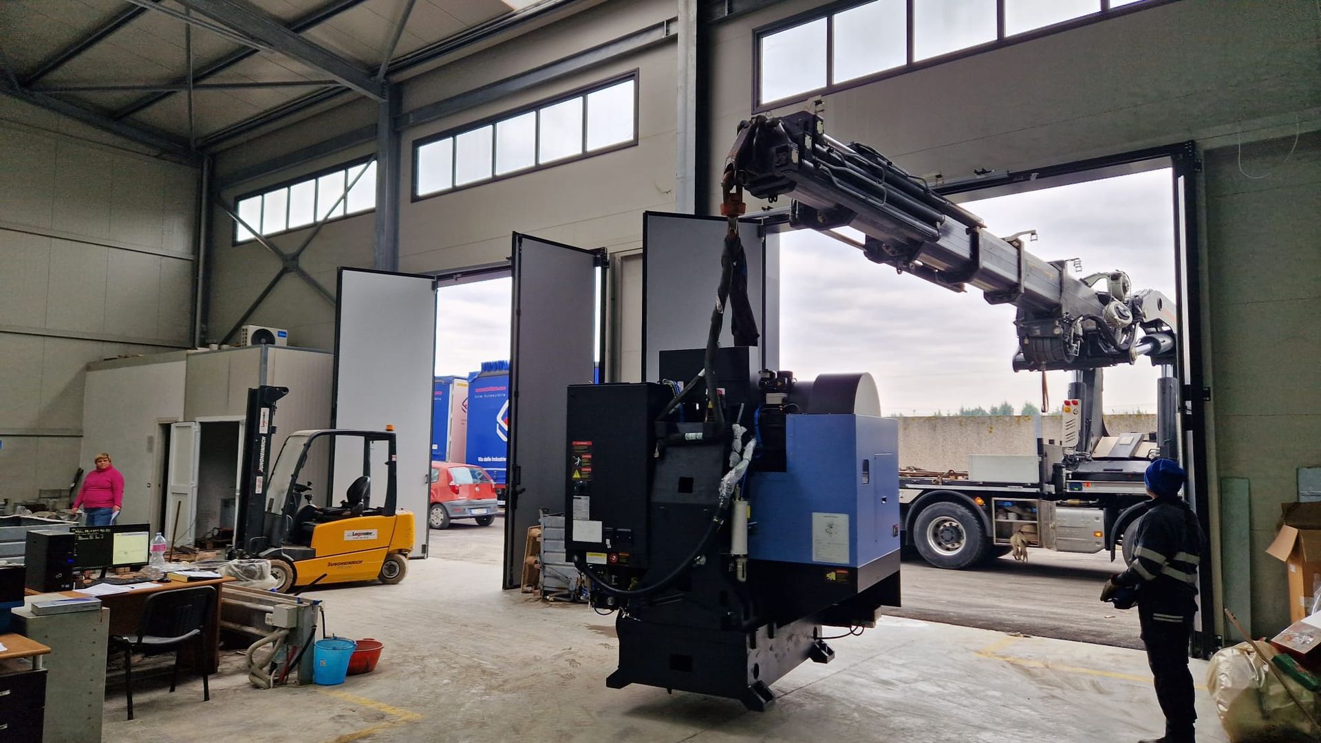 Ampia officina industriale con un veicolo gru nero-blu, attrezzi e un camion vicino a un portone di garage aperto.
