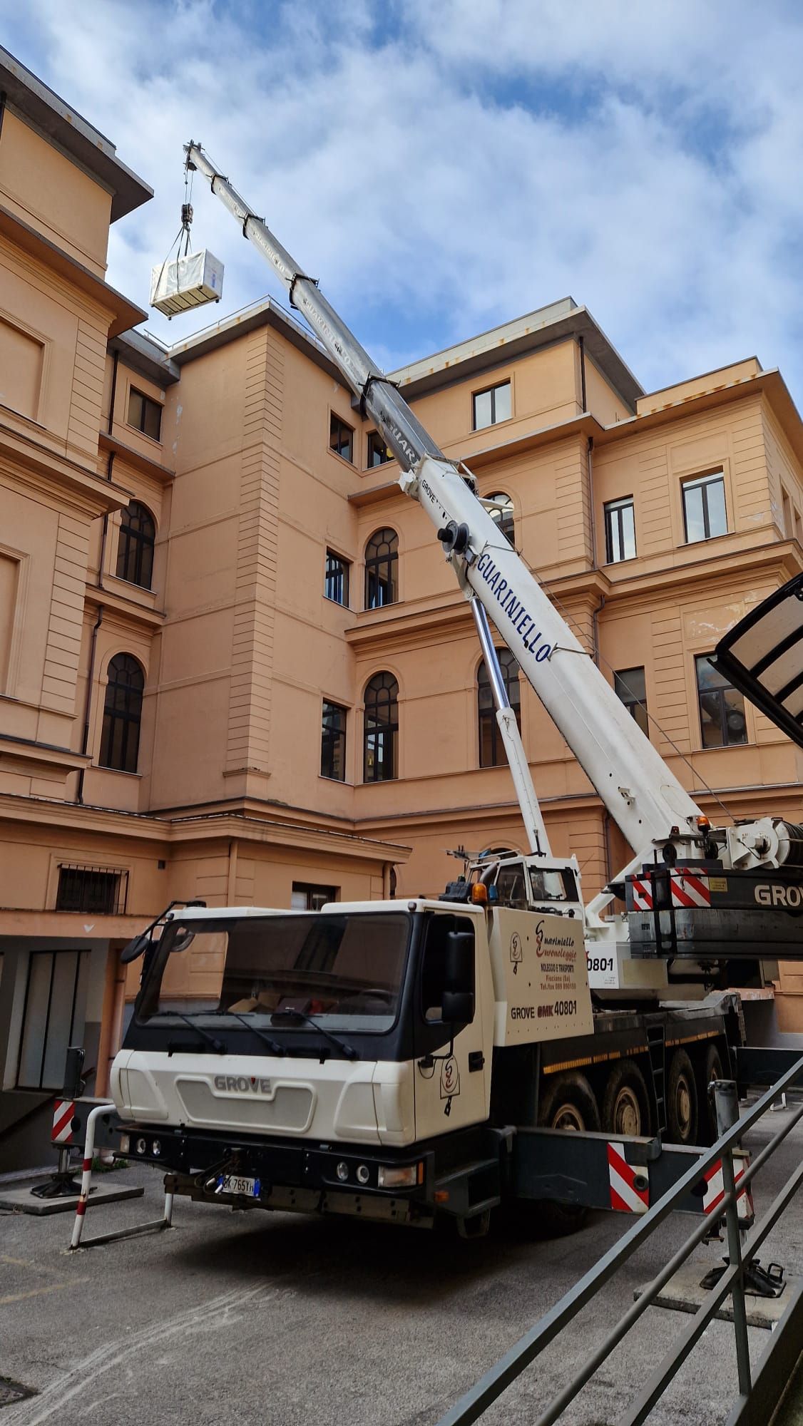 Un camion gru bianco che solleva attrezzature accanto a un edificio color pesca sotto un cielo nuvoloso.