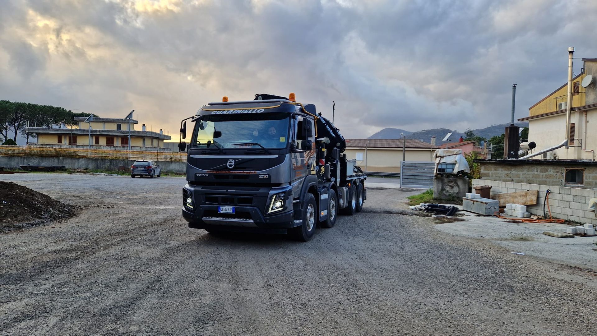 Un camion ribaltabile nero su una discarica ghiaiosa vicino ad alcuni edifici, sotto un cielo nuvoloso.