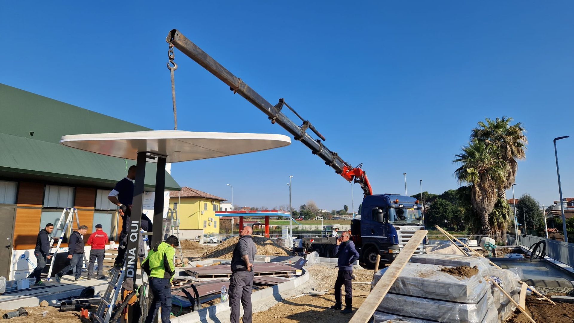 In un cantiere edile adiacente a un edificio, sotto un cielo azzurro, alcuni operai utilizzano una gru per rimuovere le macerie.