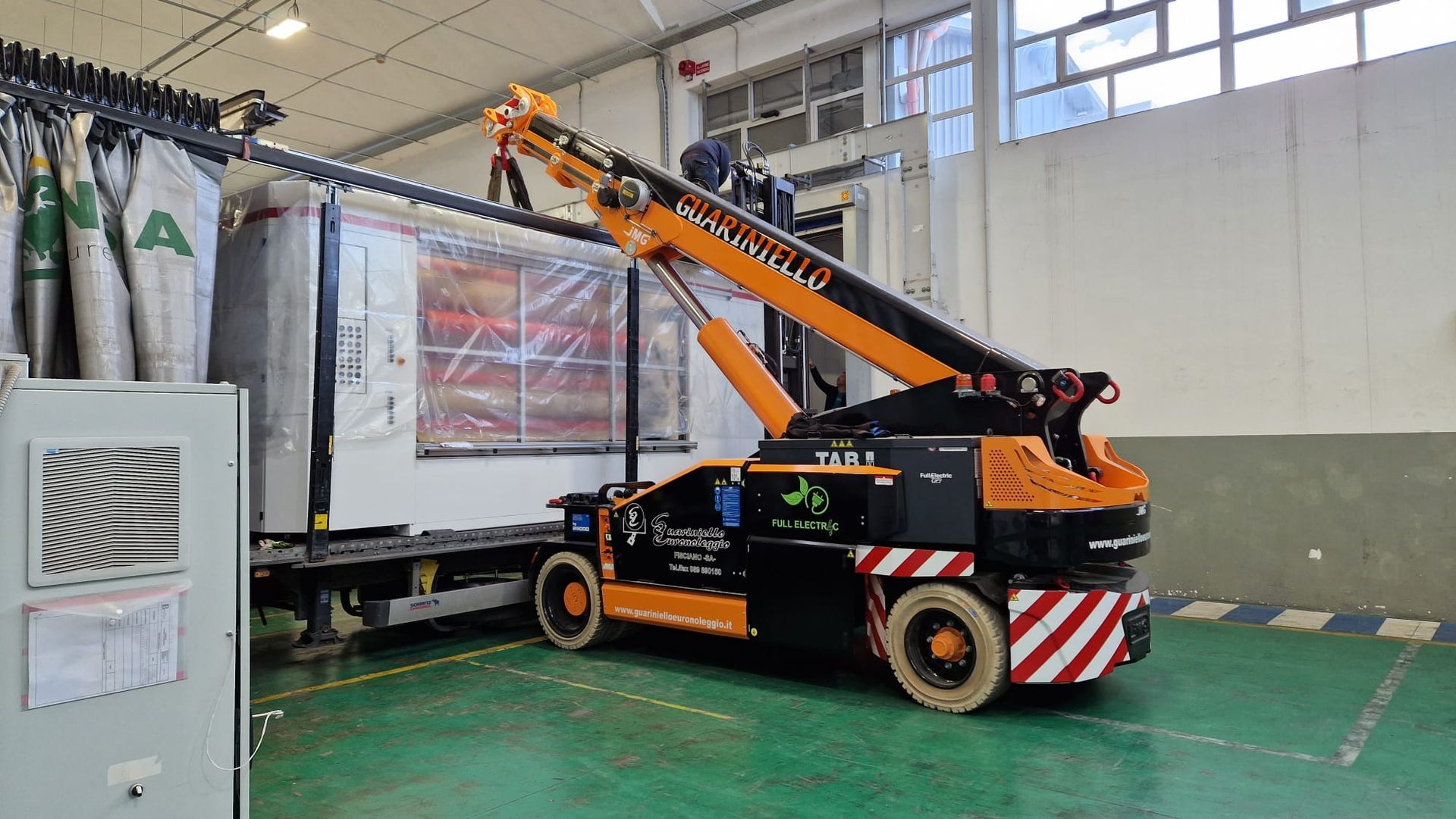 Una piattaforma aerea telescopica arancione in un magazzino, sollevata accanto a un rimorchio bianco per camion con merci imballate.