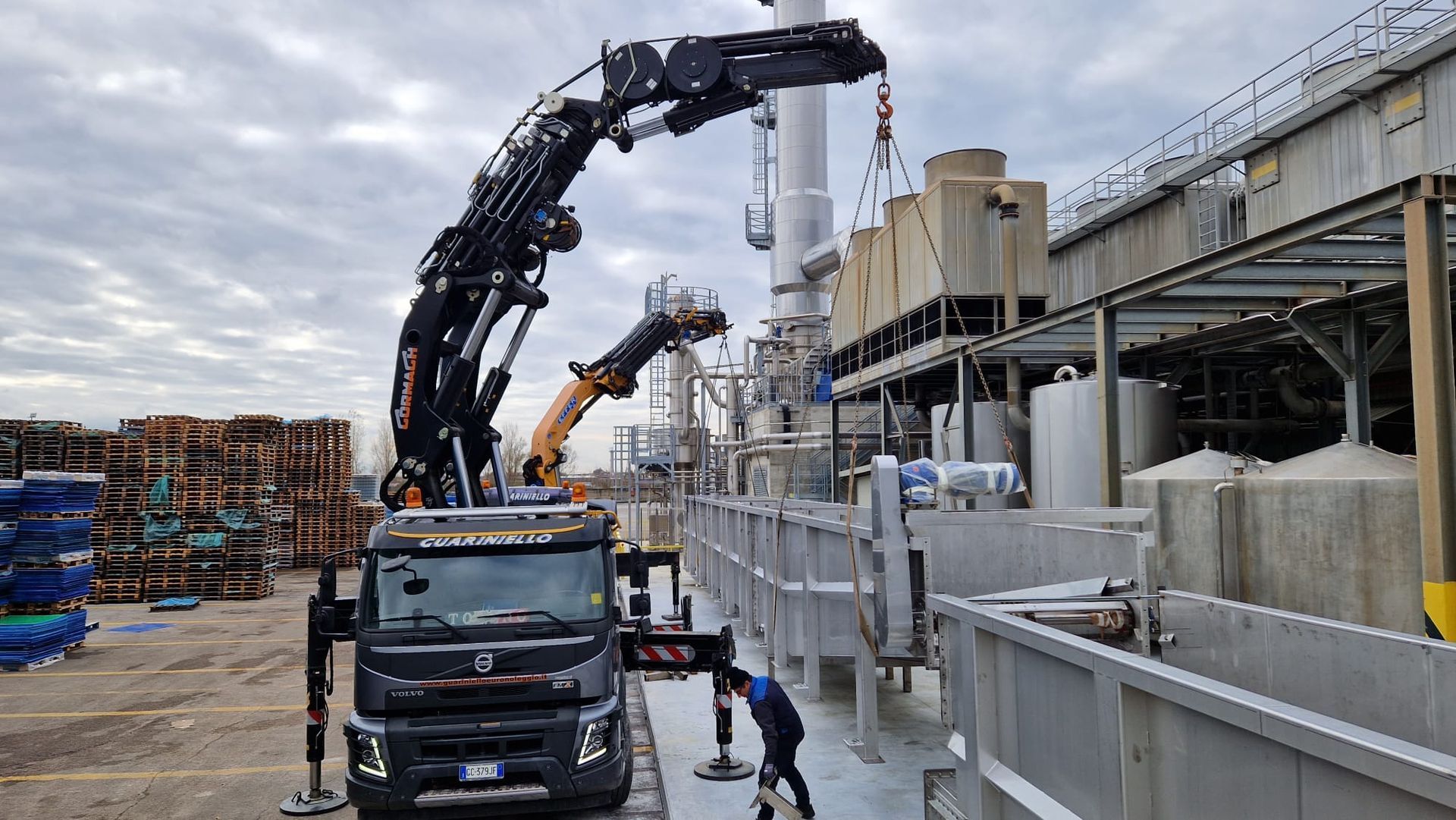Sito industriale con un camion gru che solleva detriti accanto a un edificio bianco adibito a fabbrica e pallet impilati.