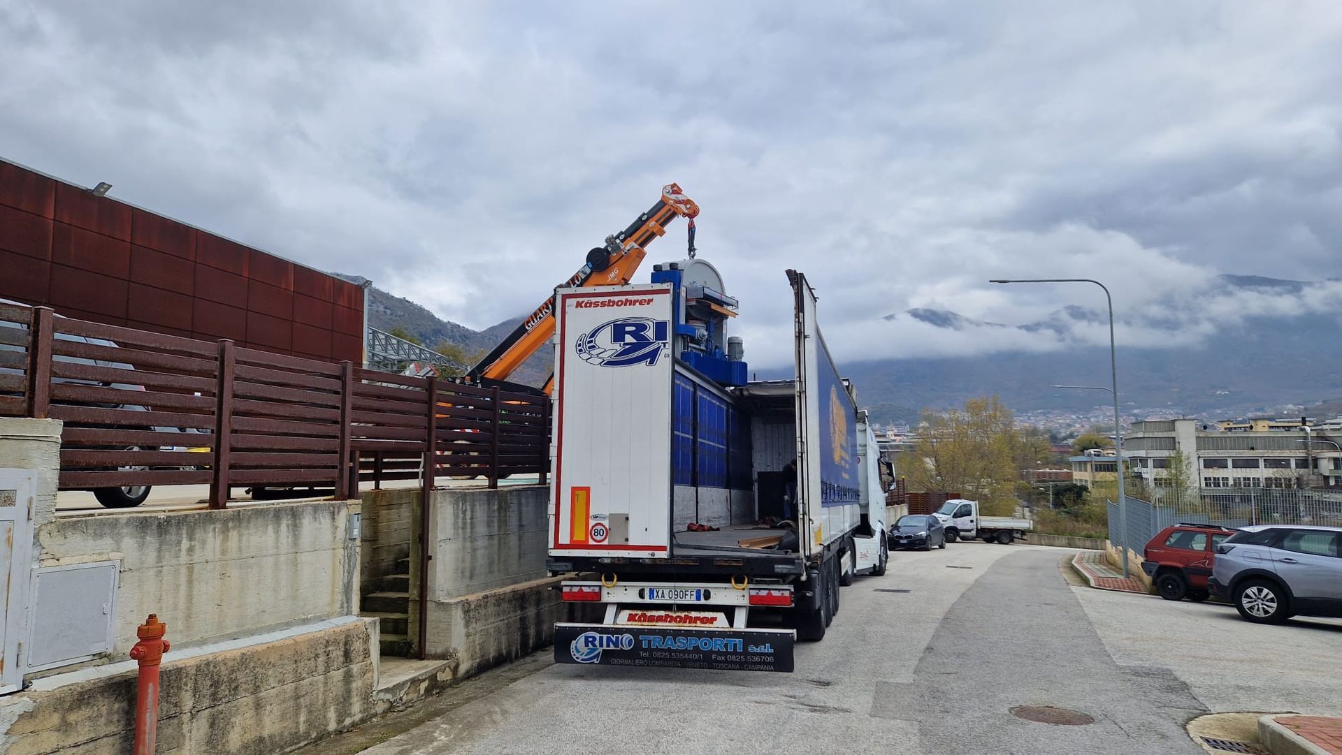 Un camioncino bianco parcheggiato su una strada stretta accanto a un muro, con montagne e cielo nuvoloso sullo sfondo.