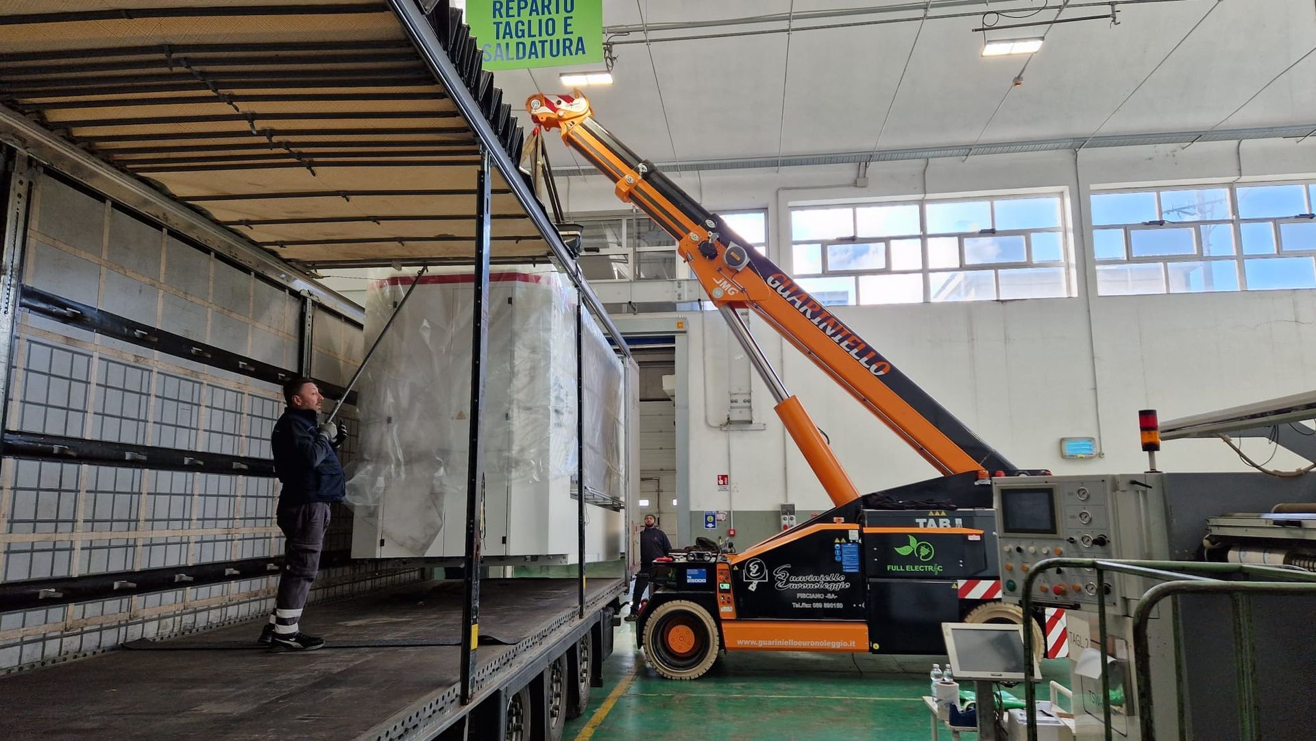 Un carrello elevatore arancione carica una grande cassa bianca su un camion, mentre un operaio la guida all'interno di un magazzino.
