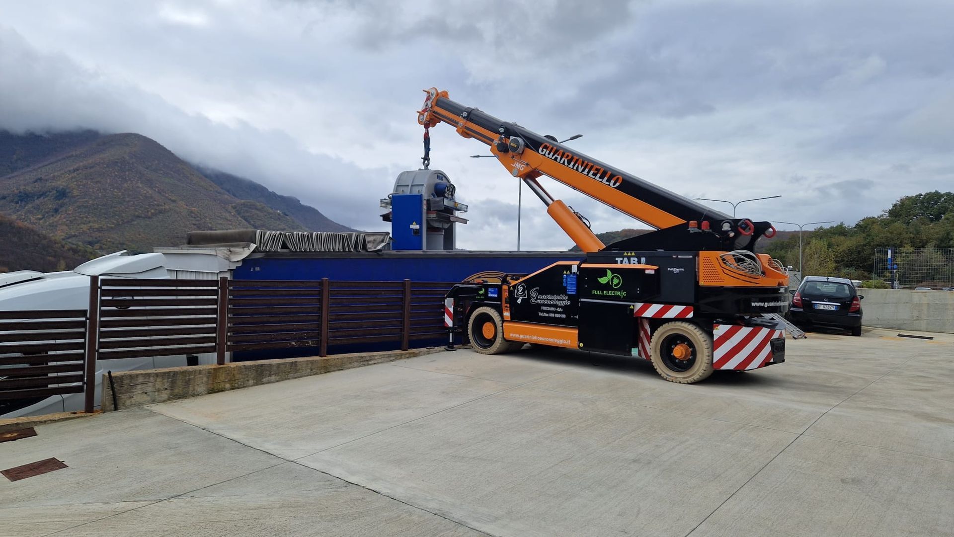 Un camion gru arancione che solleva attrezzature accanto a una struttura blu in un cantiere di montagna.