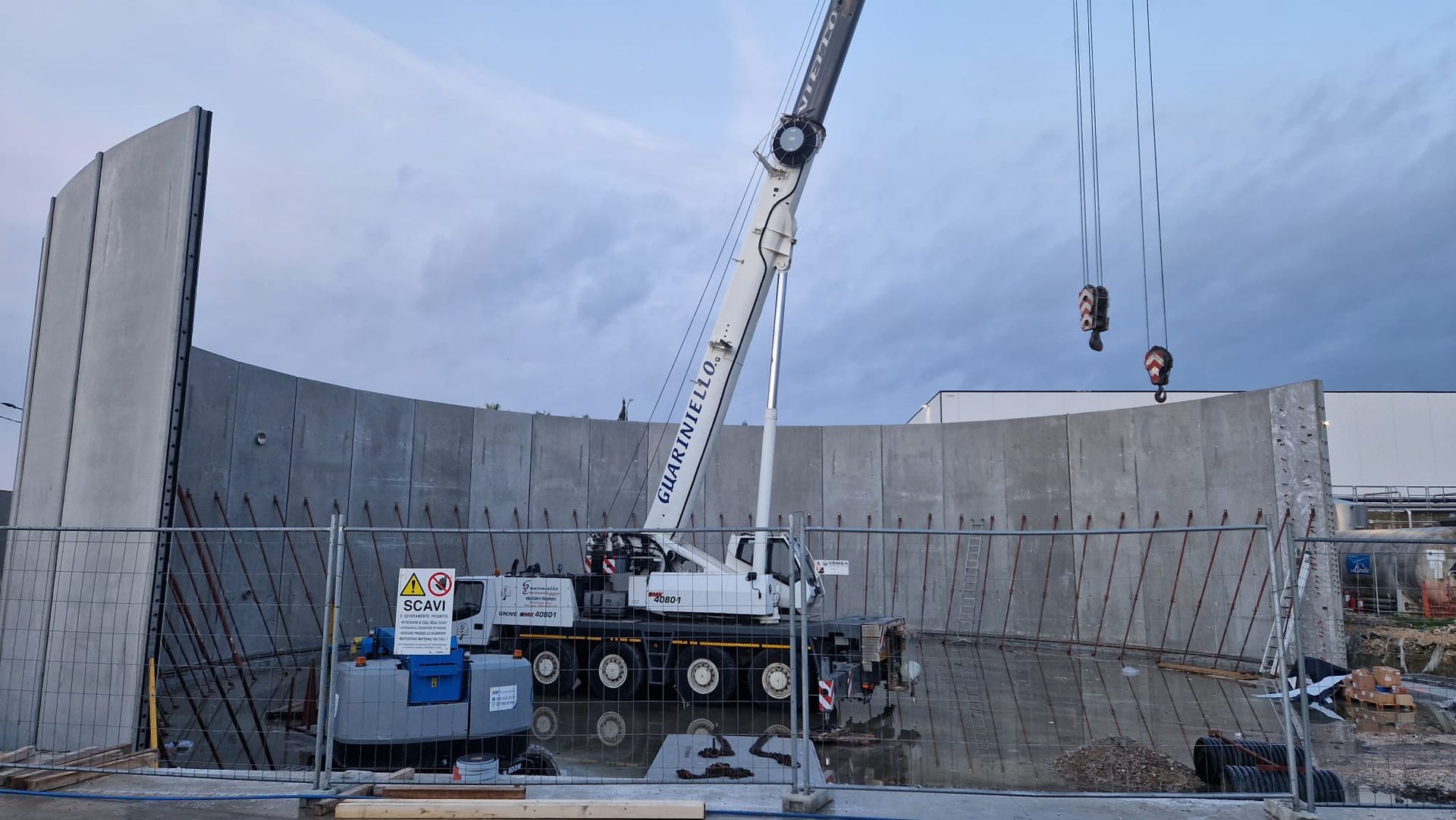 Cantiere edile con gru che solleva un segmento di muro in cemento sotto un cielo nuvoloso.