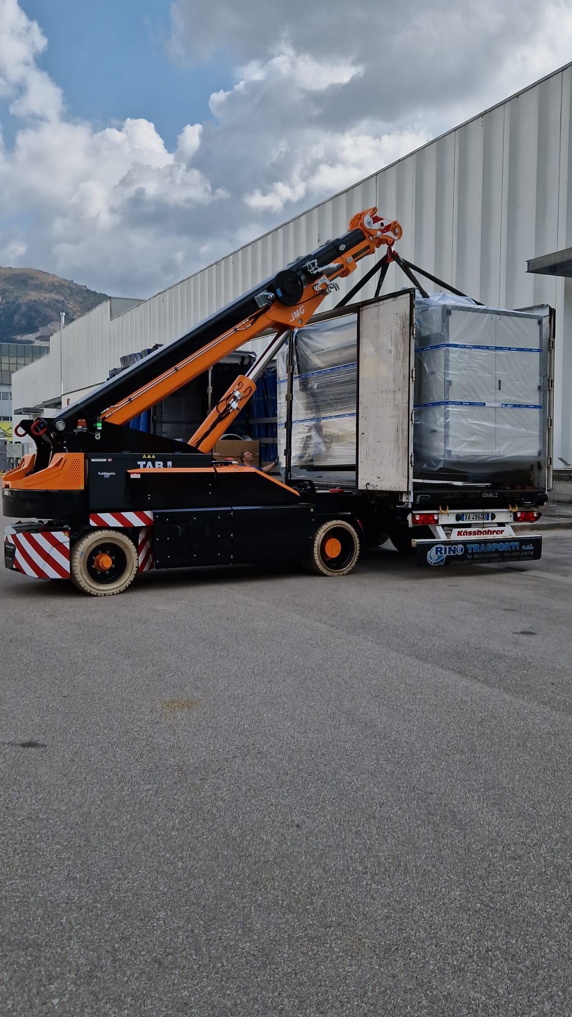 Un carrello elevatore arancione e nero carica pallet imballati accanto a un magazzino.