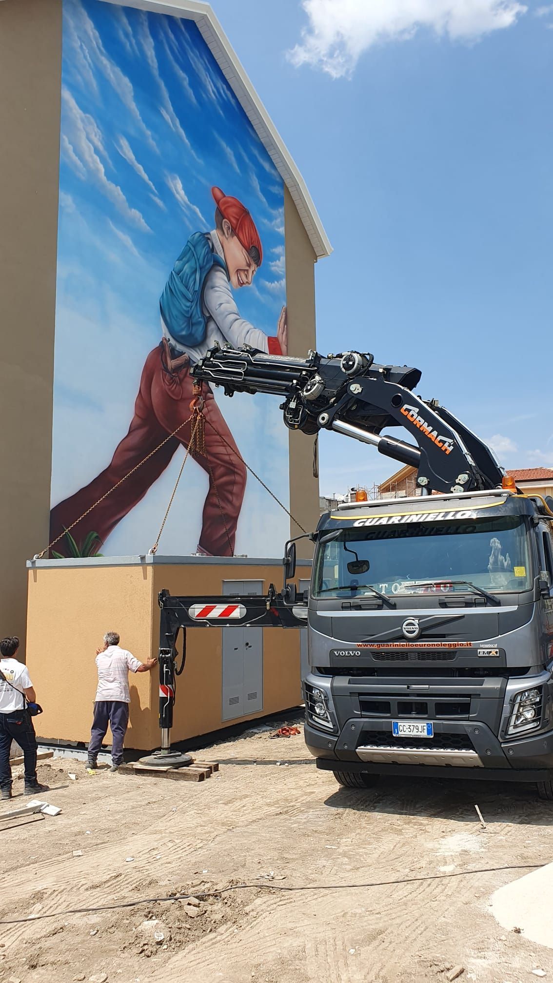 Una gru solleva dei materiali accanto a un camion e a un murale raffigurante una persona che dipinge un muro in un cantiere soleggiato.