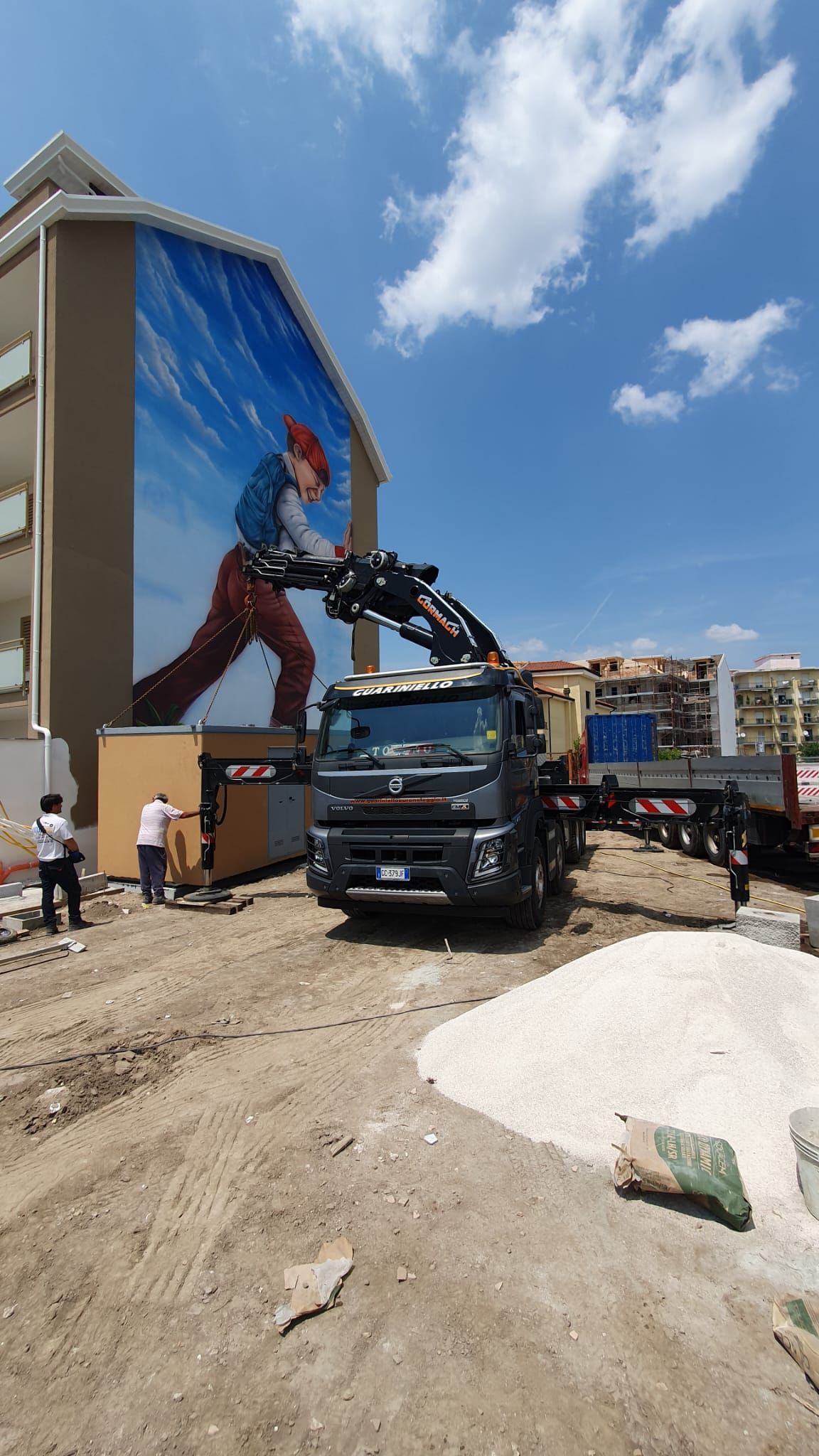 Una gru da costruzione solleva materiali accanto a un murale raffigurante un operaio che si arrampica sotto un cielo azzurro.
