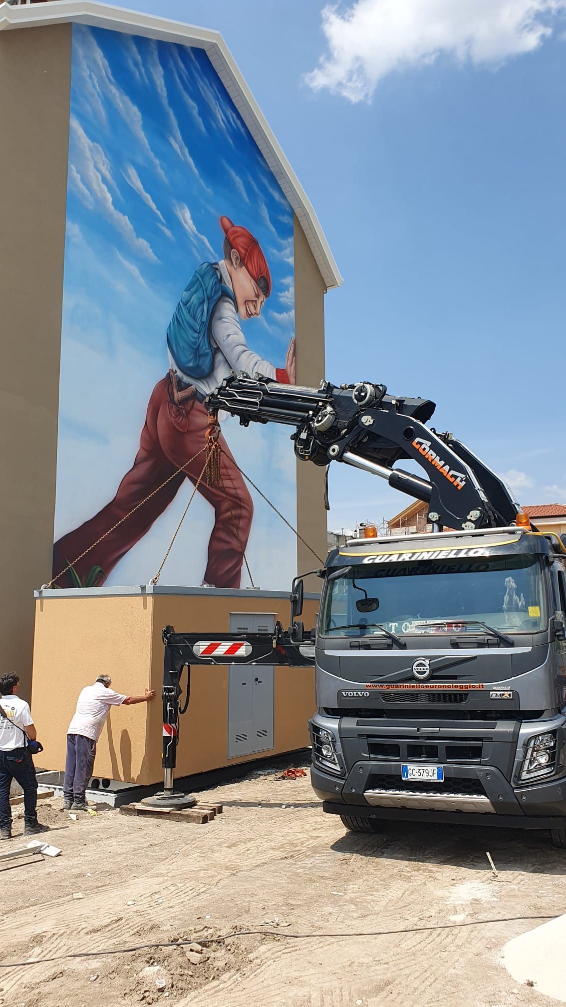 Operai e una gru su camion installano un grande murale sulla parete di un edificio sotto un cielo azzurro.