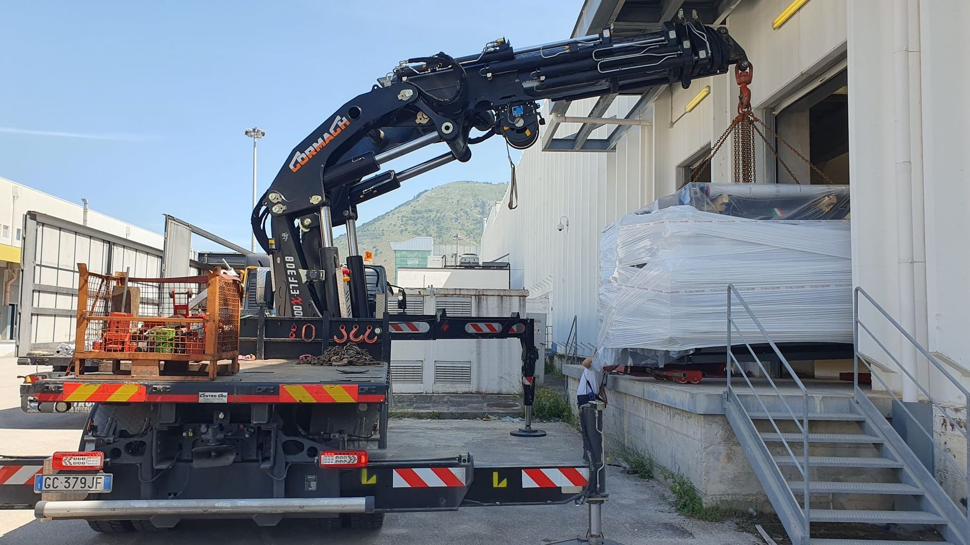 Un camion con gru scarica materiali accanto a una banchina di carico e a una scala di un magazzino.