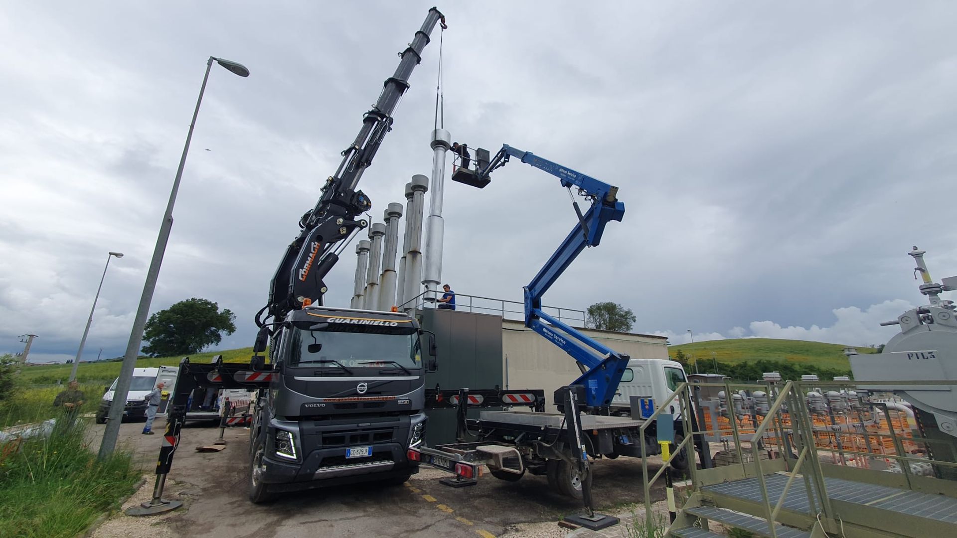 Un camion con gru e un sollevatore elettrico blu operano in un cantiere stradale sotto un cielo nuvoloso.
